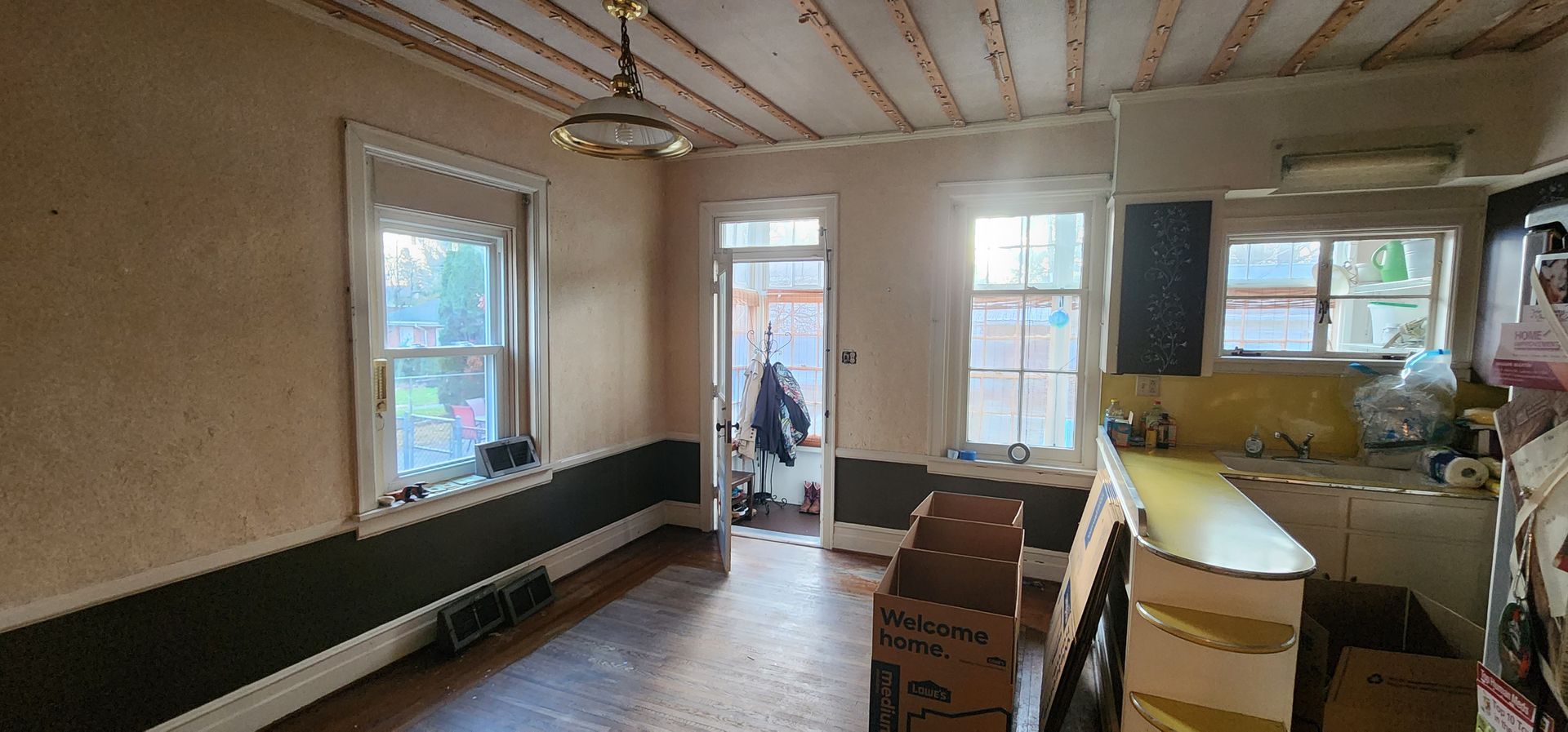 A room under renovation with beige walls, a dark grey lower border, exposed ceiling rafters, and kitchen area.