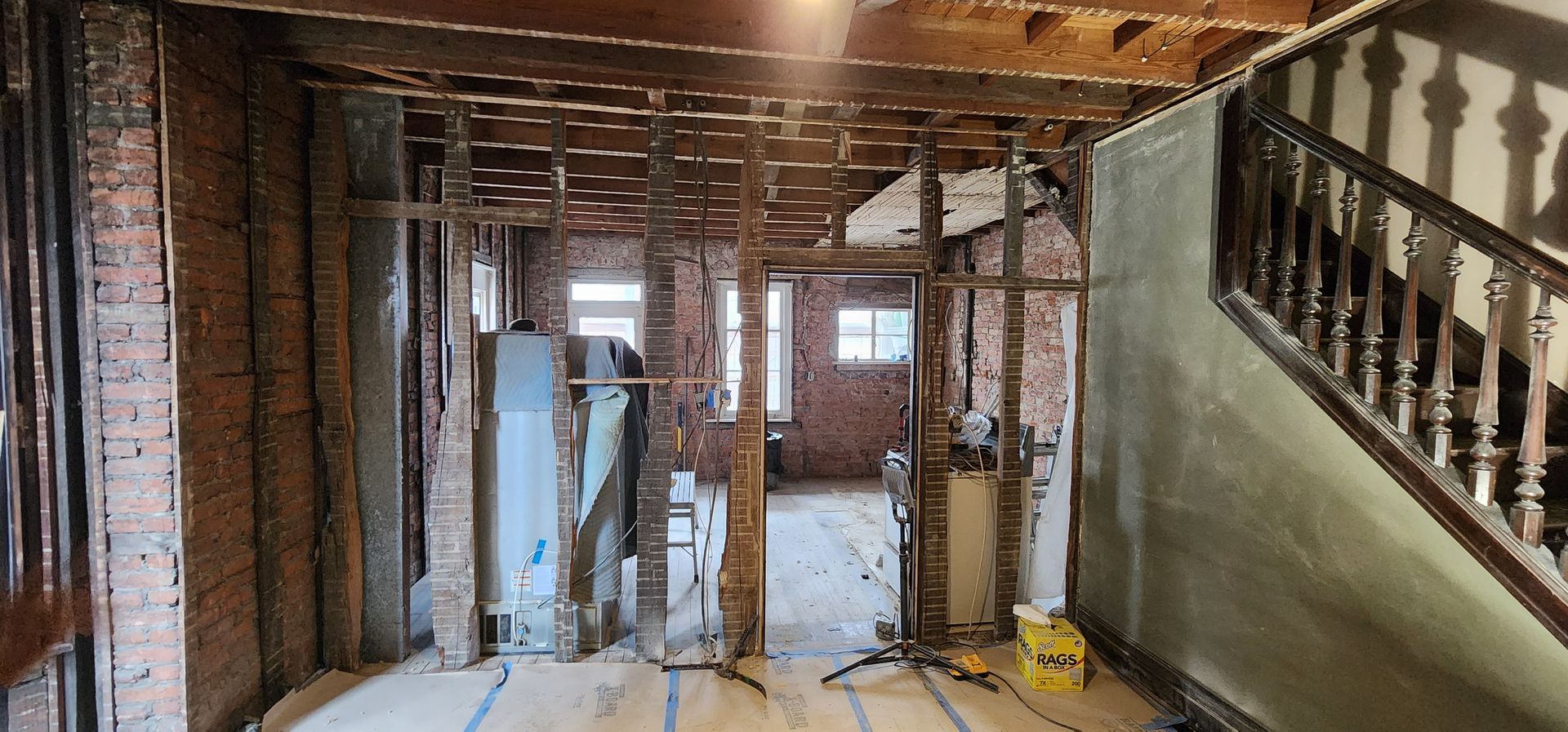 A room under renovation with exposed brick walls, wooden ceiling joists, a water heater, and a wooden staircase.