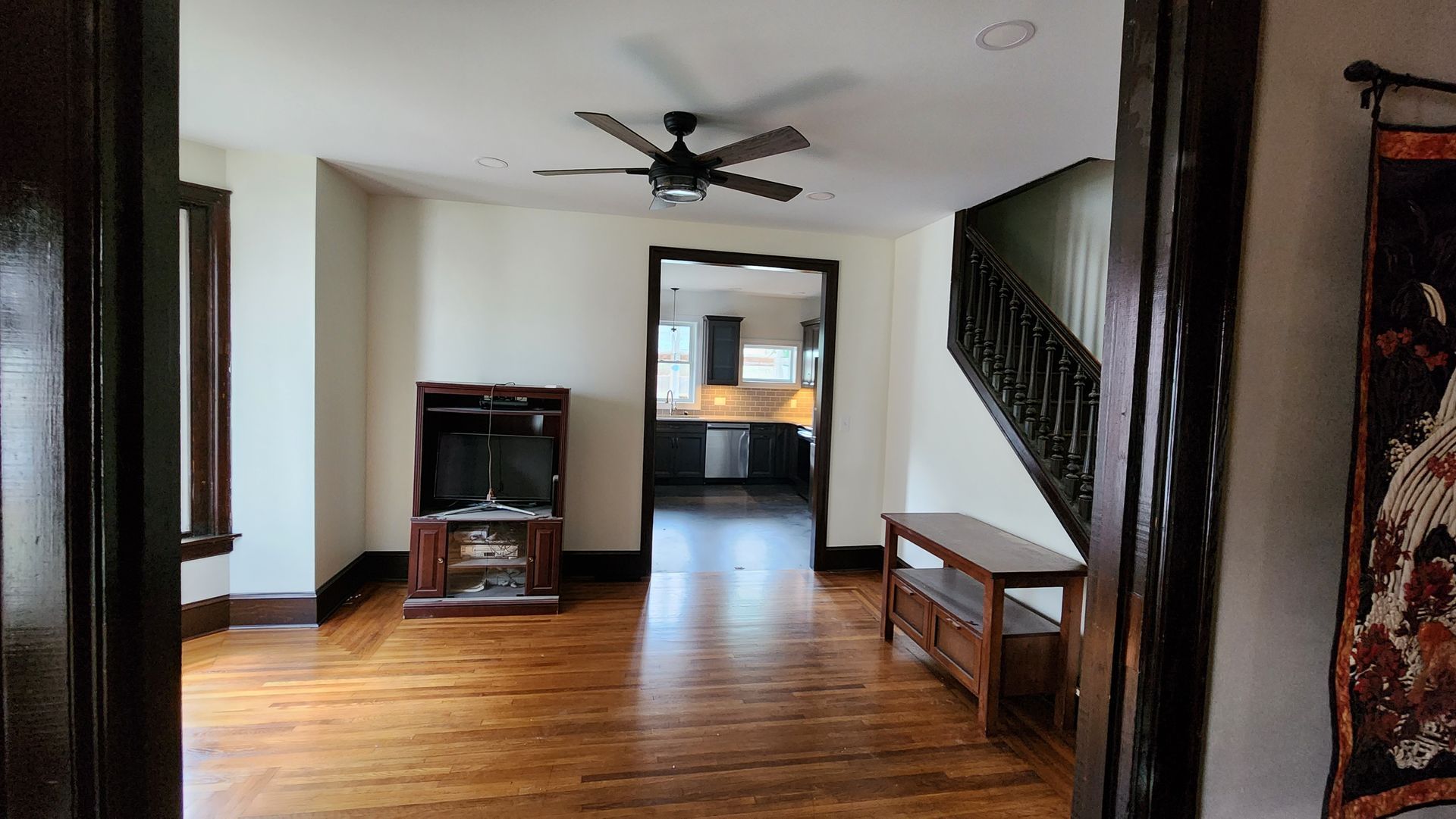 A spacious room with wood floors leads through a doorway to a kitchen. A staircase with a dark railing sits to the right.