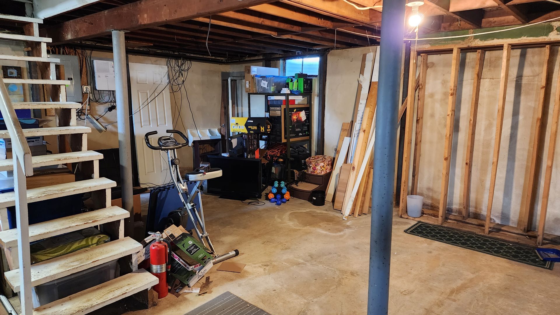 A cluttered basement with a wooden staircase on the left, an exercise bike, stored equipment, and unfinished wall framing.