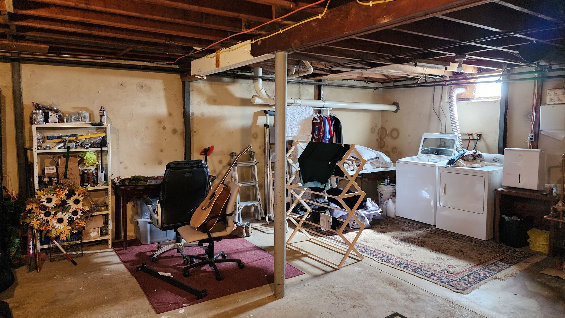 A cluttered unfinished basement with a desk, guitar, drying rack, and laundry machines under exposed ceiling joists.