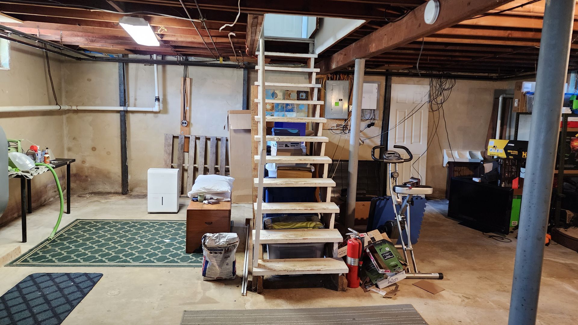 An unfinished basement with concrete floors, white walls, exposed wooden ceiling beams, and wooden stairs in the center.
