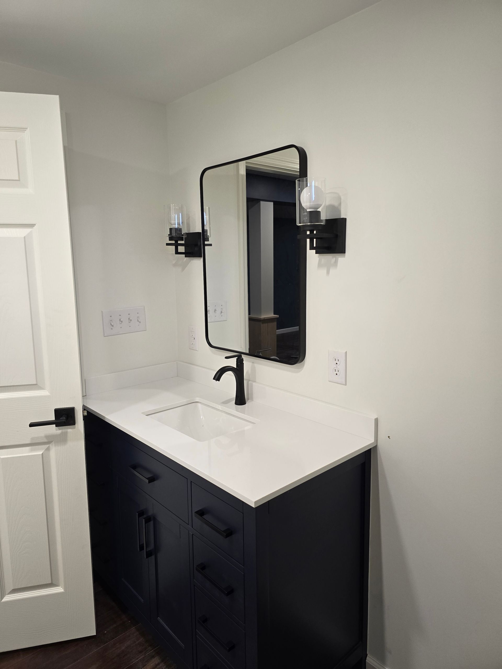 A modern vanity with a dark blue cabinet, white countertop, black faucet, rectangular mirror, and two wall sconces.