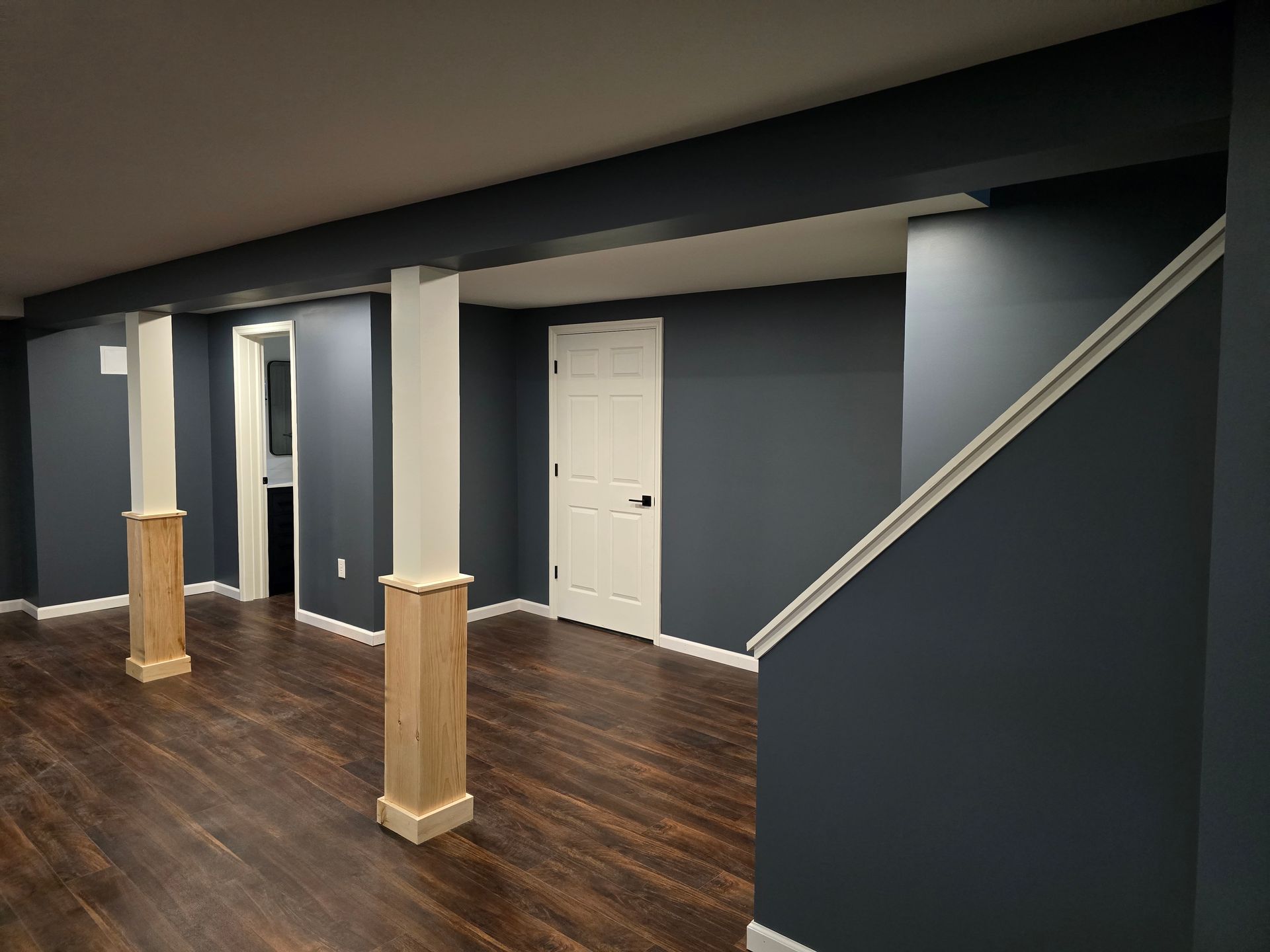 A renovated basement with dark blue walls, dark wood floors, a white door, and two pillars with wood-trimmed bases.