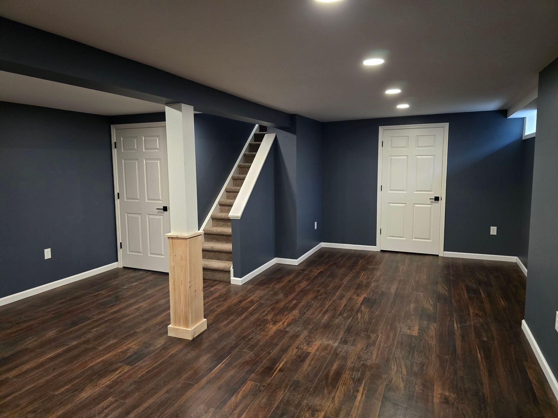 A finished basement with dark blue walls, dark wood floors, a wooden support pillar, stairs, and two white doors.