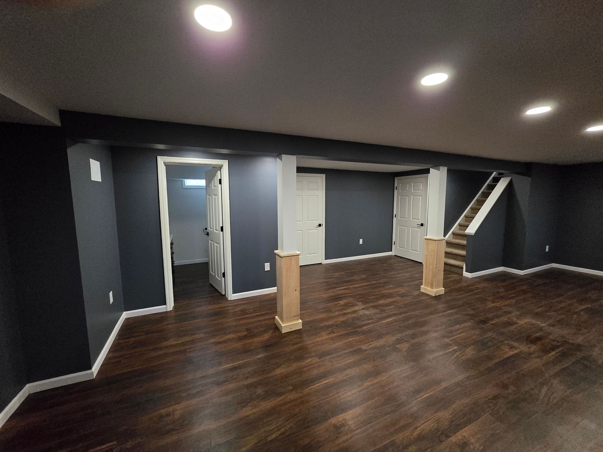 A finished basement featuring dark blue walls, dark hardwood floors, two wooden support posts, and a staircase.