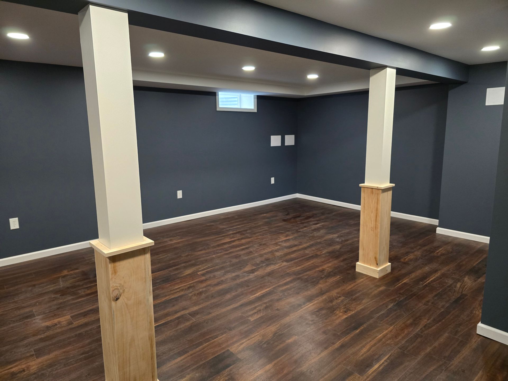 A finished basement featuring dark blue walls, dark wood floors, and two support columns with light wood bases.