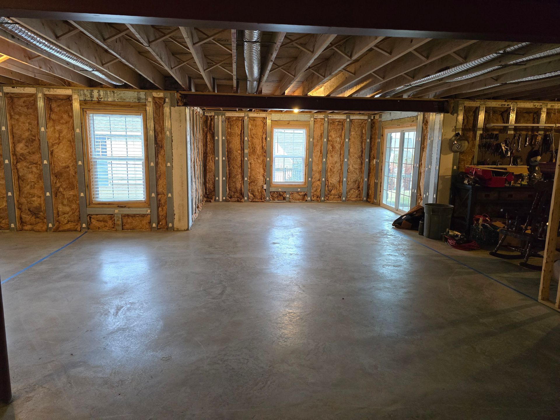A basement interior with a concrete floor, exposed ceiling joists, steel wall framing, two windows, and a sliding door.