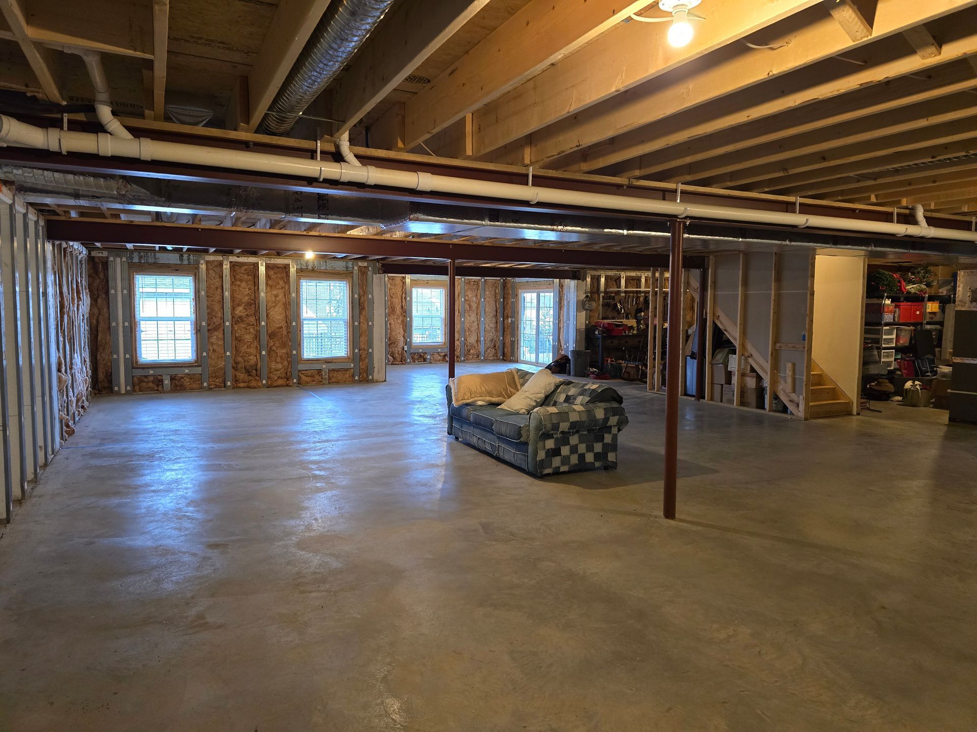 A large, unfinished basement with concrete floors, exposed ceiling joists, stud walls, and a patterned couch in the center.
