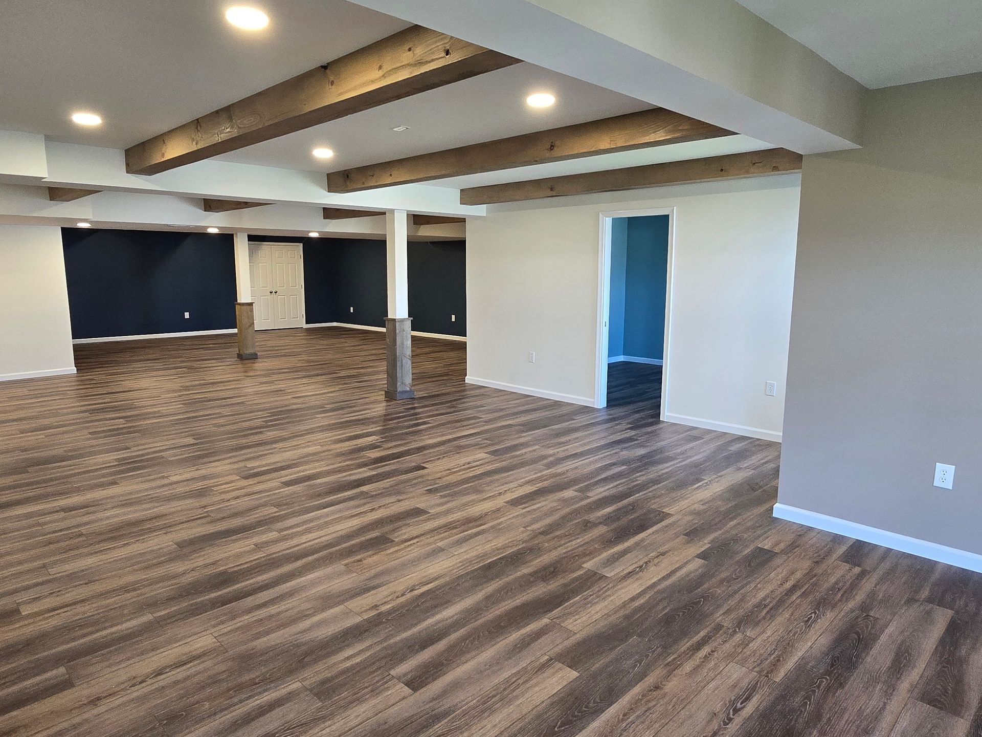 A basement interior with wood-look flooring, exposed wooden ceiling beams, neutral walls, and a dark blue accent wall.
