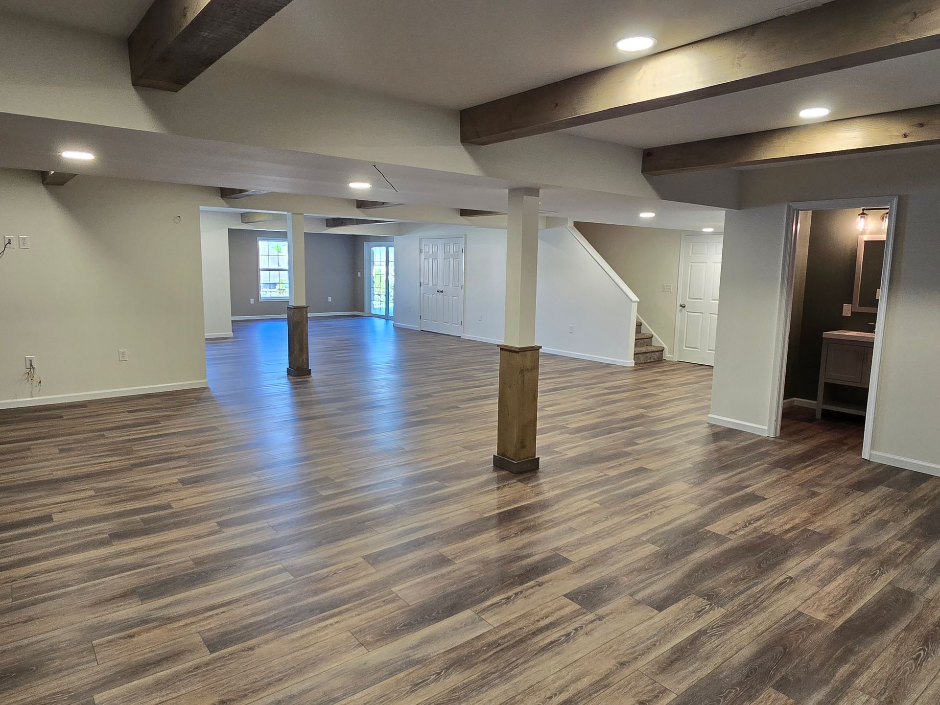 A spacious basement with wood-look flooring, light gray walls, dark wood support beams and posts, and recessed lighting.