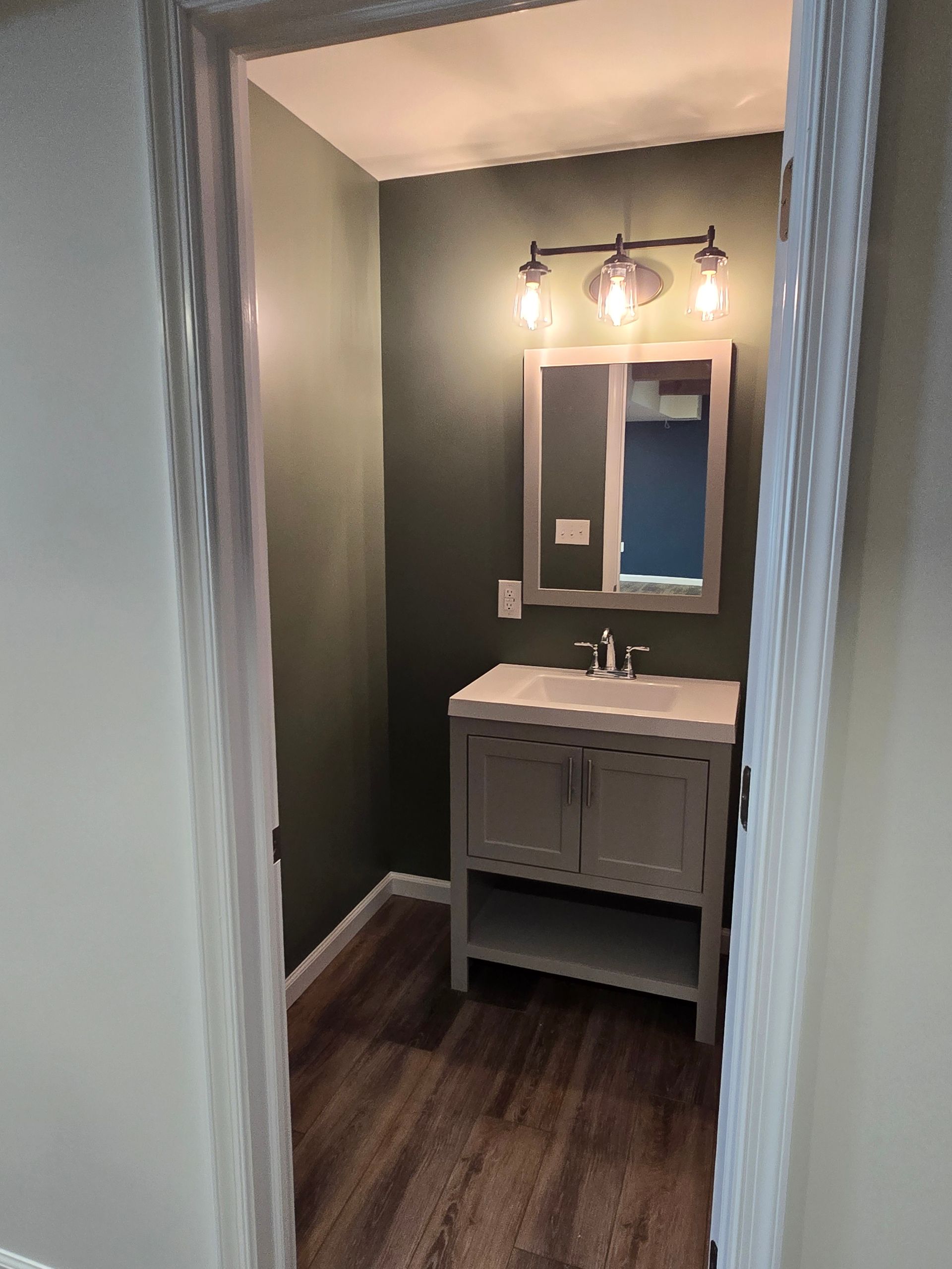A small bathroom with dark green walls, a gray vanity cabinet, a rectangular mirror, and wood-look flooring.