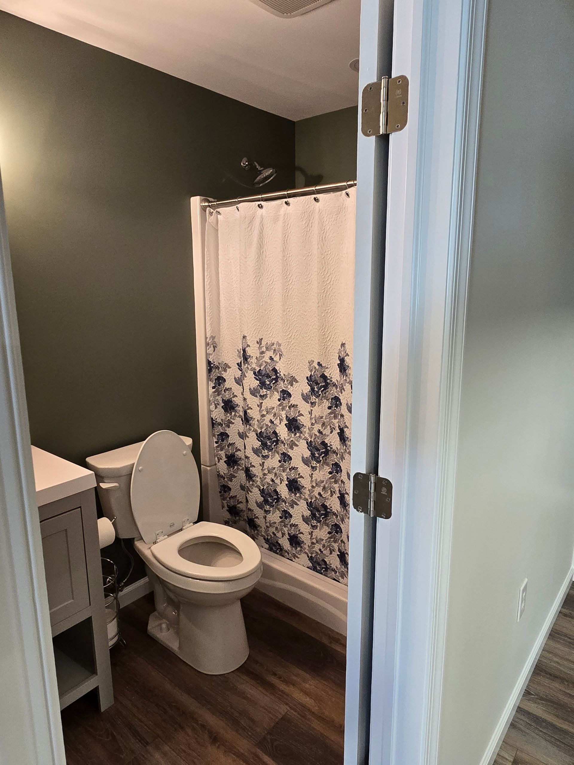 A small bathroom with dark green walls, a white toilet, a floral shower curtain, and a gray vanity cabinet.