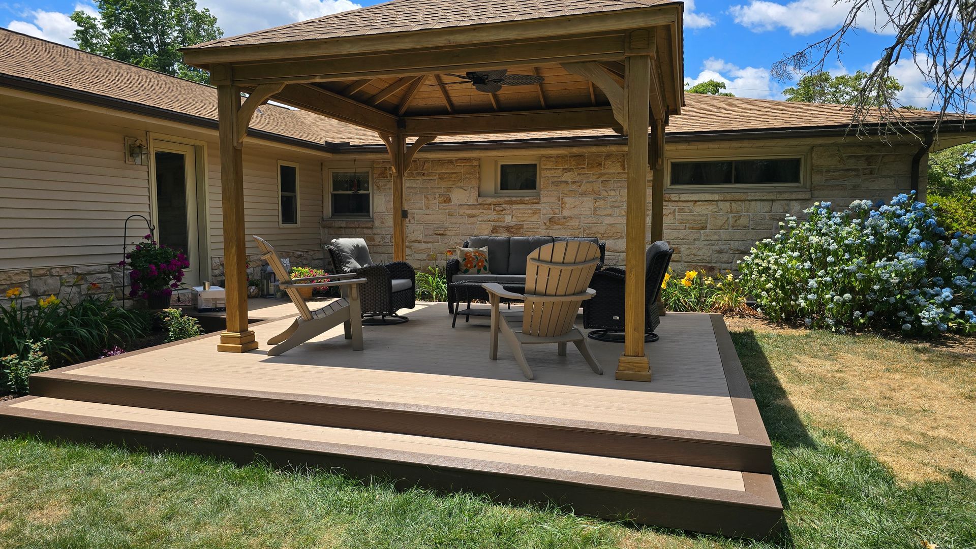 A wooden gazebo on a raised backyard patio with outdoor furniture, stone house siding, and lush garden plants.
