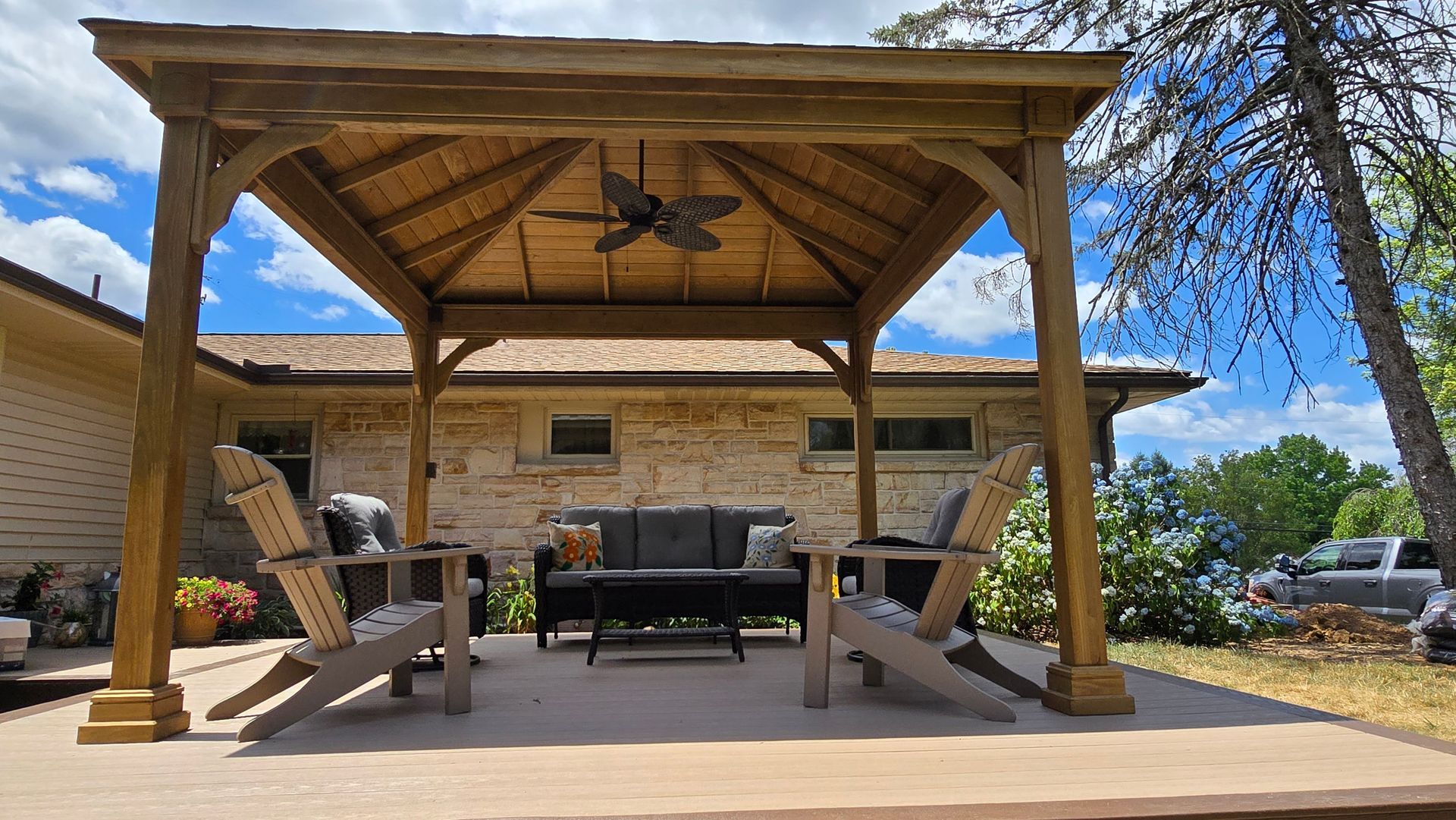 A wood gazebo covers an outdoor patio set with chairs and a sofa on a stone porch next to a house under a blue sky.
