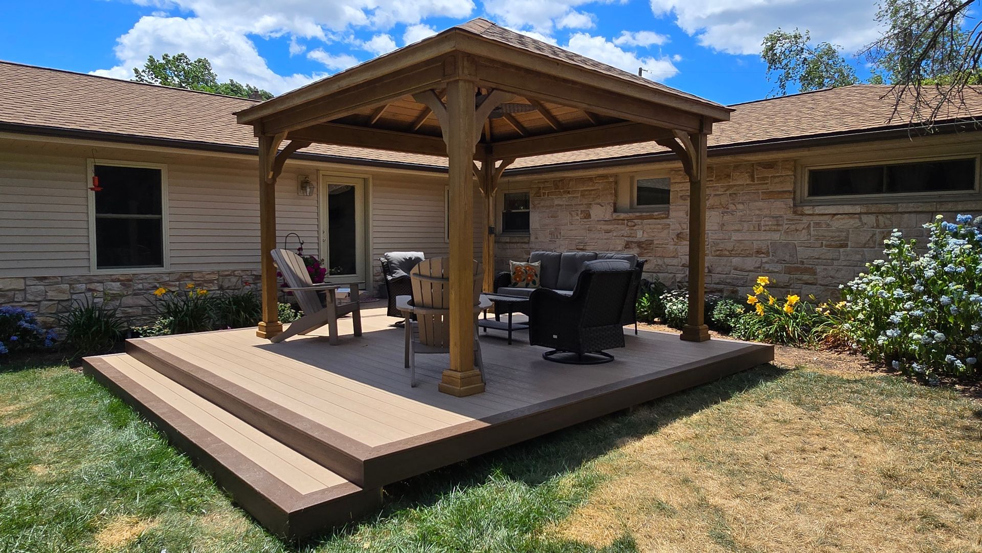 A wooden gazebo sits on a multi-level deck behind a home with stone accents and patio furniture in a sunny backyard.