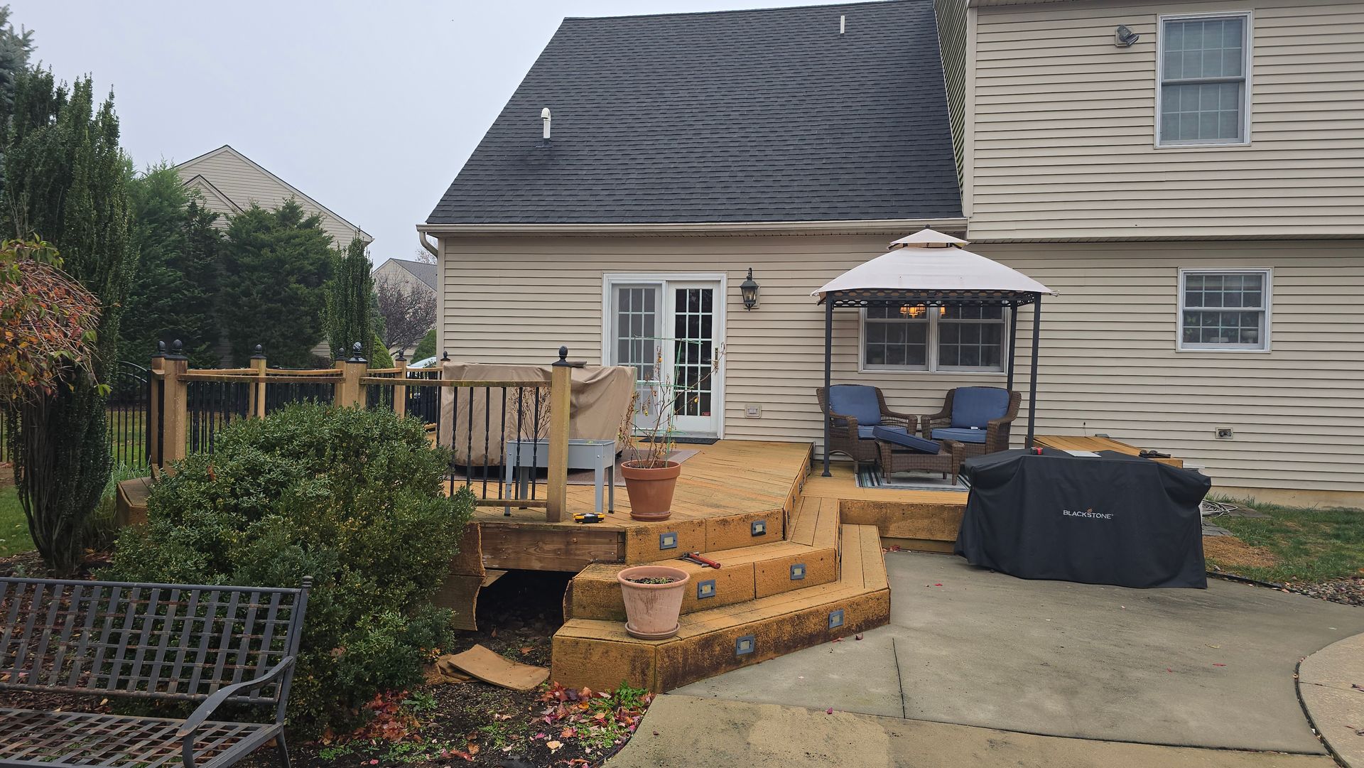 A multi-level wood deck with a gazebo, patio furniture, and potted plants attached to the back of a tan siding house.