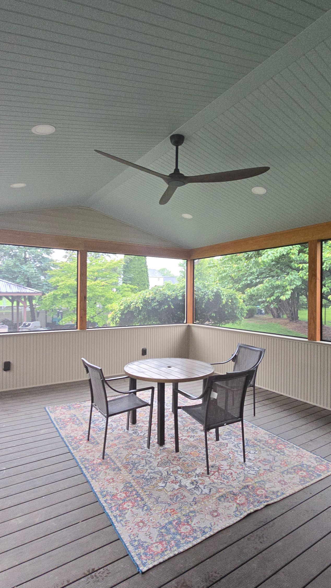 A covered porch with a ceiling fan, table, and chairs arranged on a patterned rug, overlooking a lush green yard.