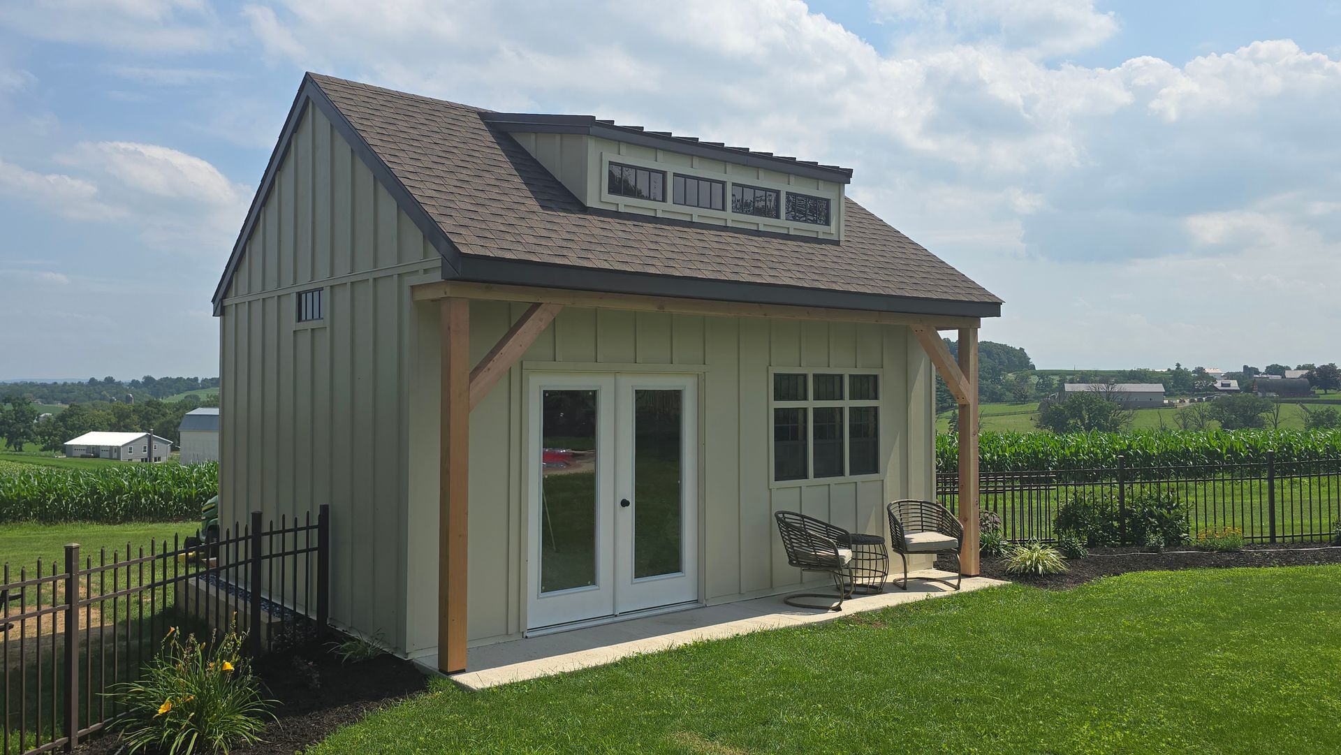 A beige, modern shed with a dormer, double glass doors, and a small porch featuring two chairs in a grassy yard.