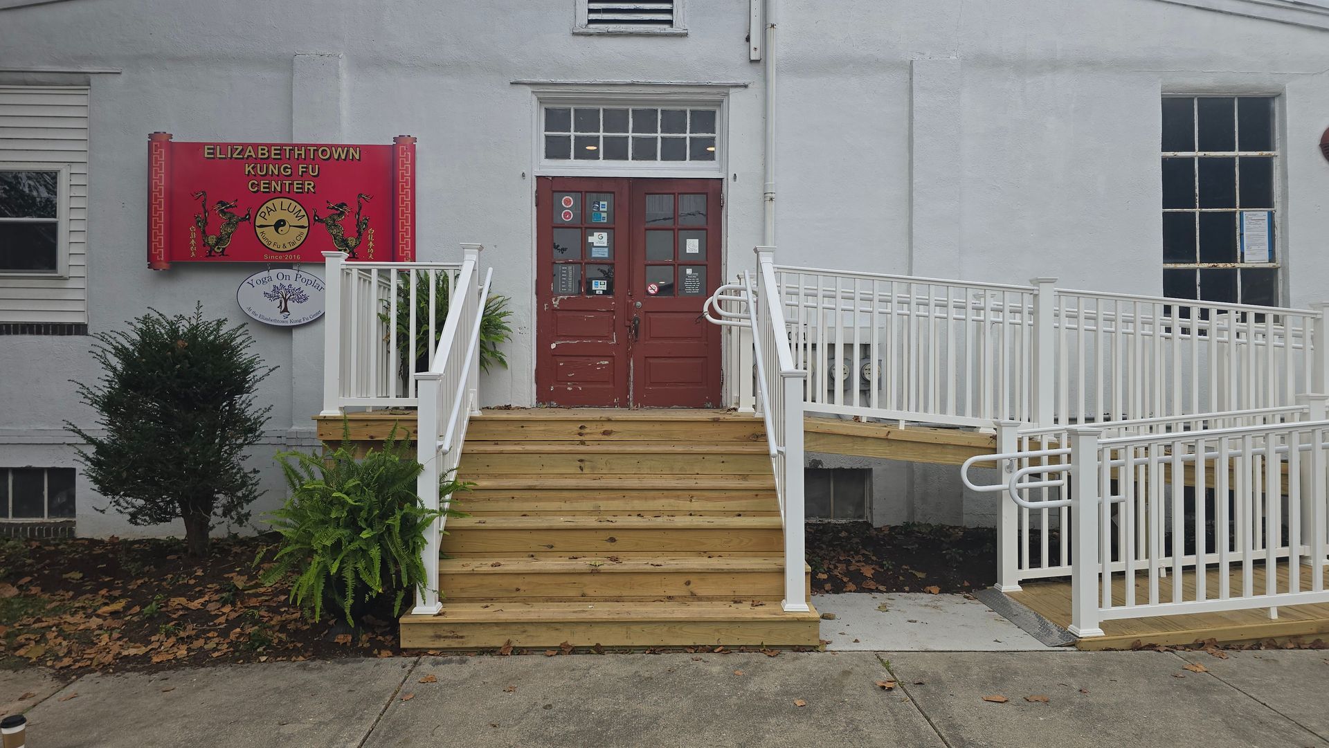 A white building entrance featuring a set of wooden stairs, a wheelchair ramp with white railings, and red double doors.