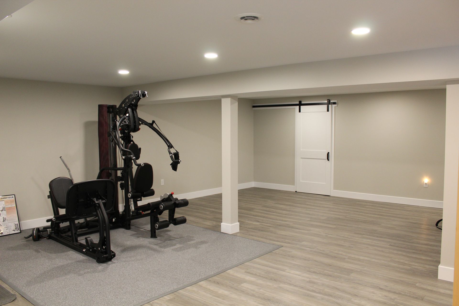 A home gym area in a basement with light gray walls, wood-look flooring, a gray rug, a weight machine, and a sliding door.