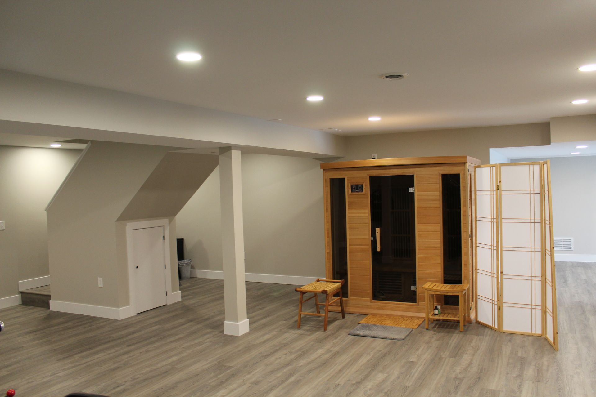 A wooden sauna with a glass door stands in a modern, neutral-toned basement with light-colored flooring and recessed lights.