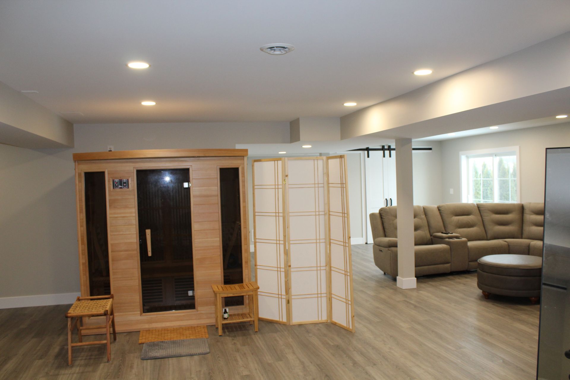 A wooden sauna, a room divider, and a beige sectional sofa in a spacious finished basement with light wood flooring.