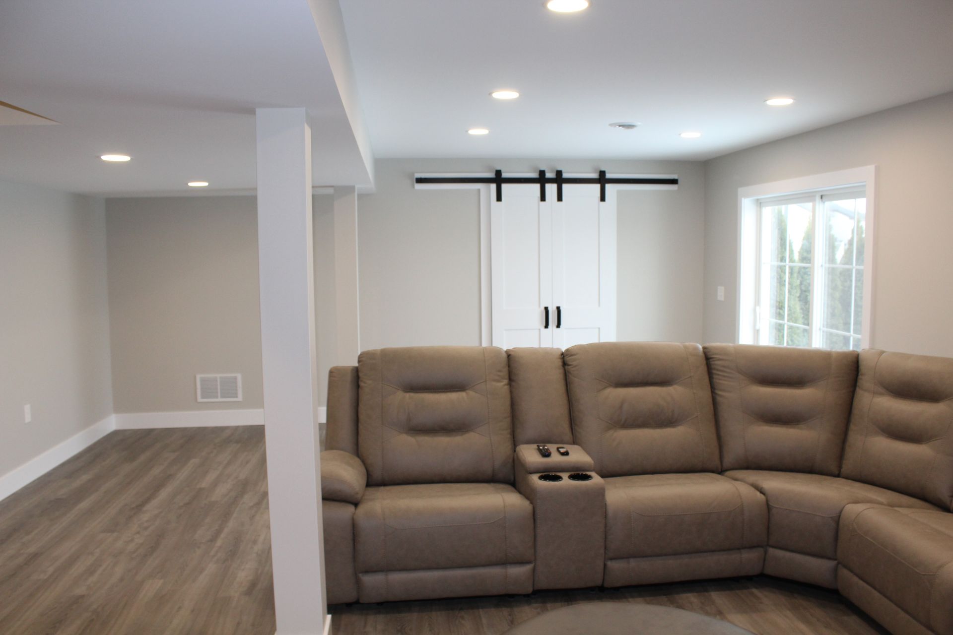 A modern basement living area features a beige sectional sofa on gray wood-look flooring near a white sliding barn door.