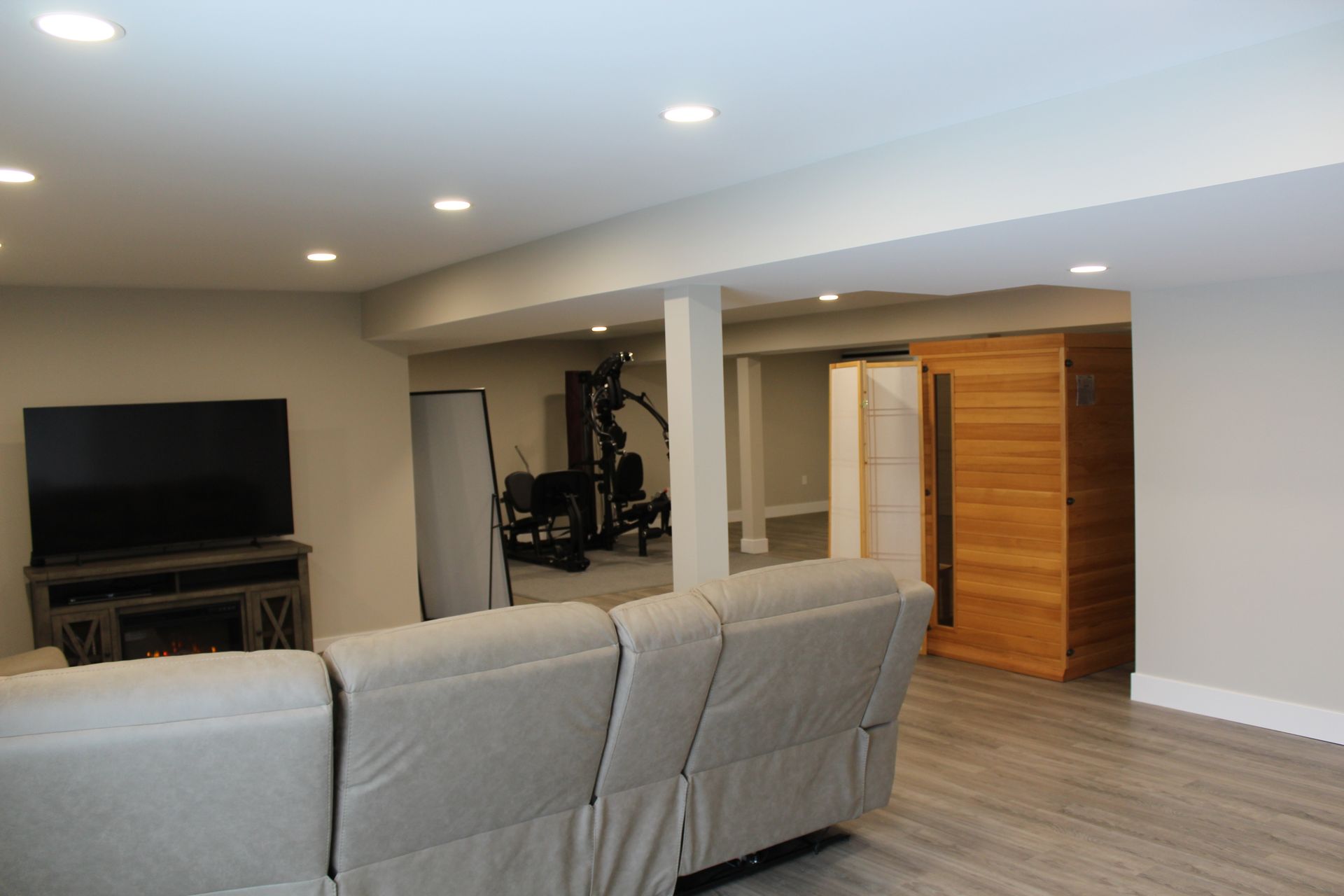 A basement with a grey sofa, a TV on a stand, a home gym in the background, and a wooden sauna in the corner.