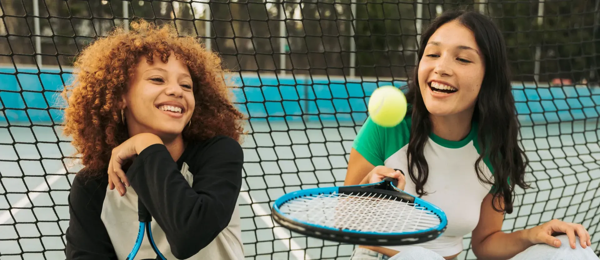 Two teen girls are laughing while bouncing a ball on a tennis racket