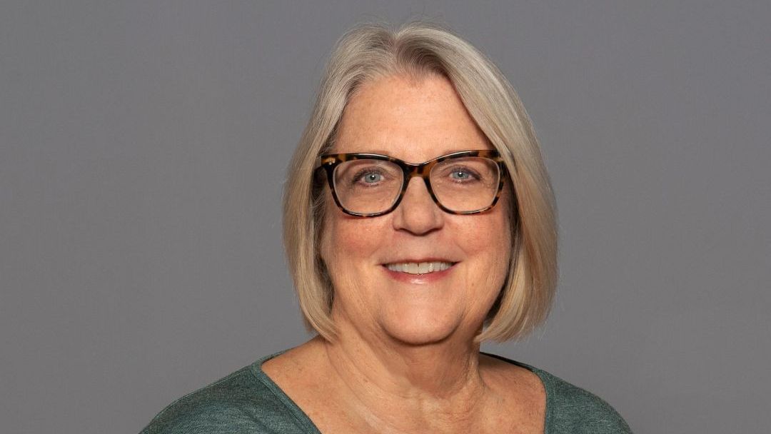 Portrait of a smiling woman with gray hair and glasses against a plain gray background