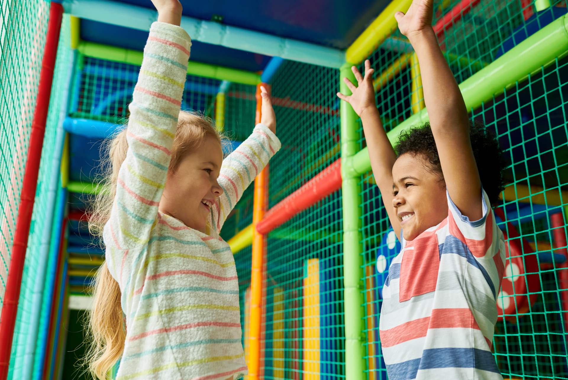 Two children in colorful play structure, raising arms, smiling.