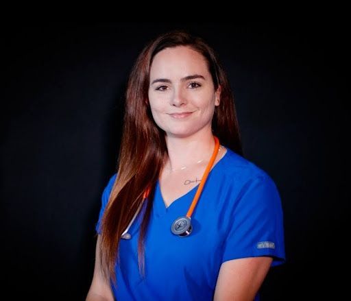 Woman in blue medical scrubs with orange trim, standing against a black background and smiling softly
