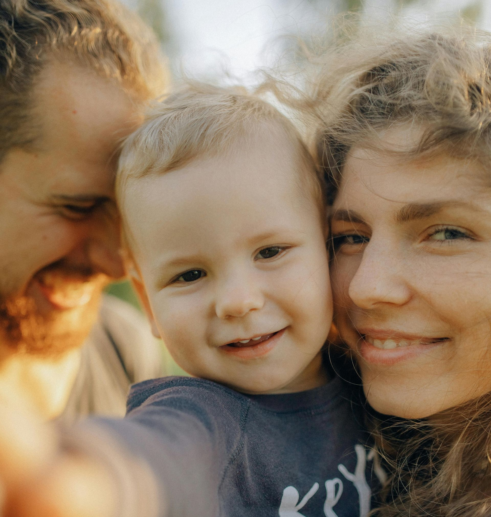 Three smiling people posing closely for a selfie outdoors in warm sunlight.