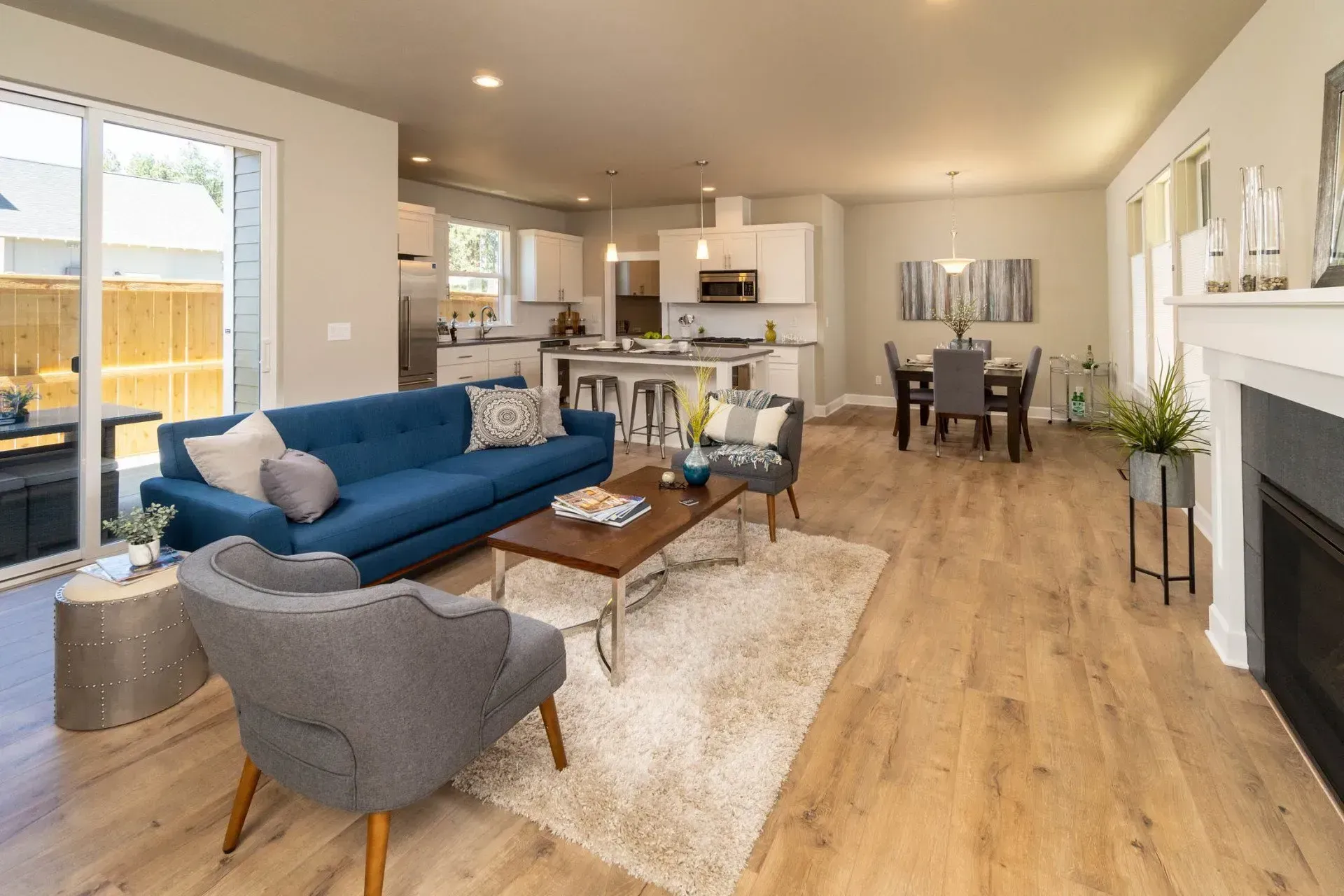 Bright open-plan living room with blue sofa, gray chair, wooden floors, and dining area in the background