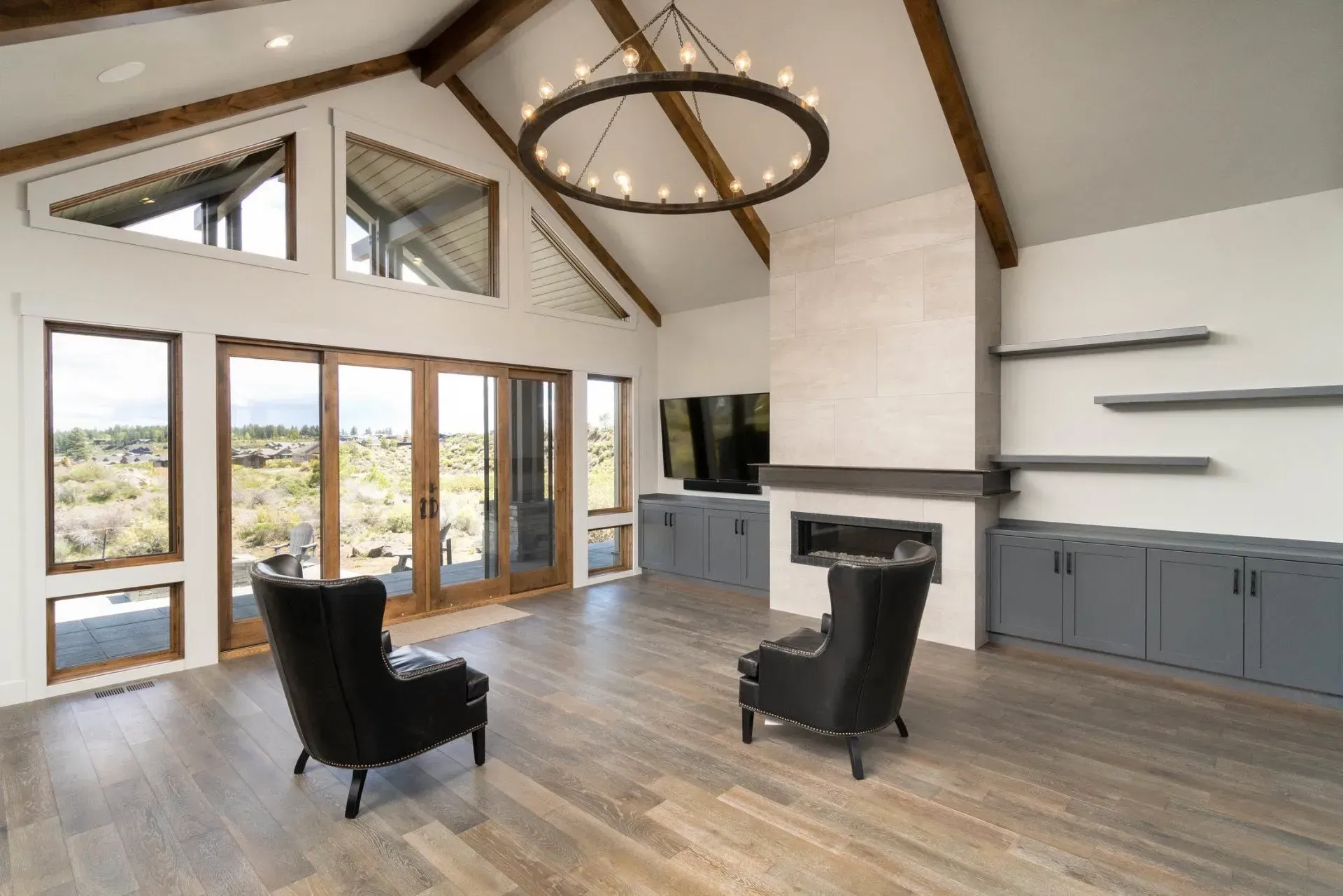 Modern living room with vaulted ceiling, glass doors, gray cabinets, and two black chairs by a fireplace.