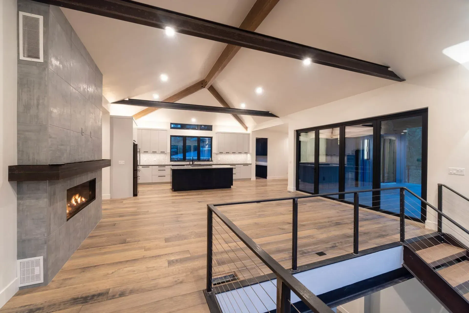 Modern open-concept living room with fireplace, exposed beams, wood floors, and large sliding glass doors