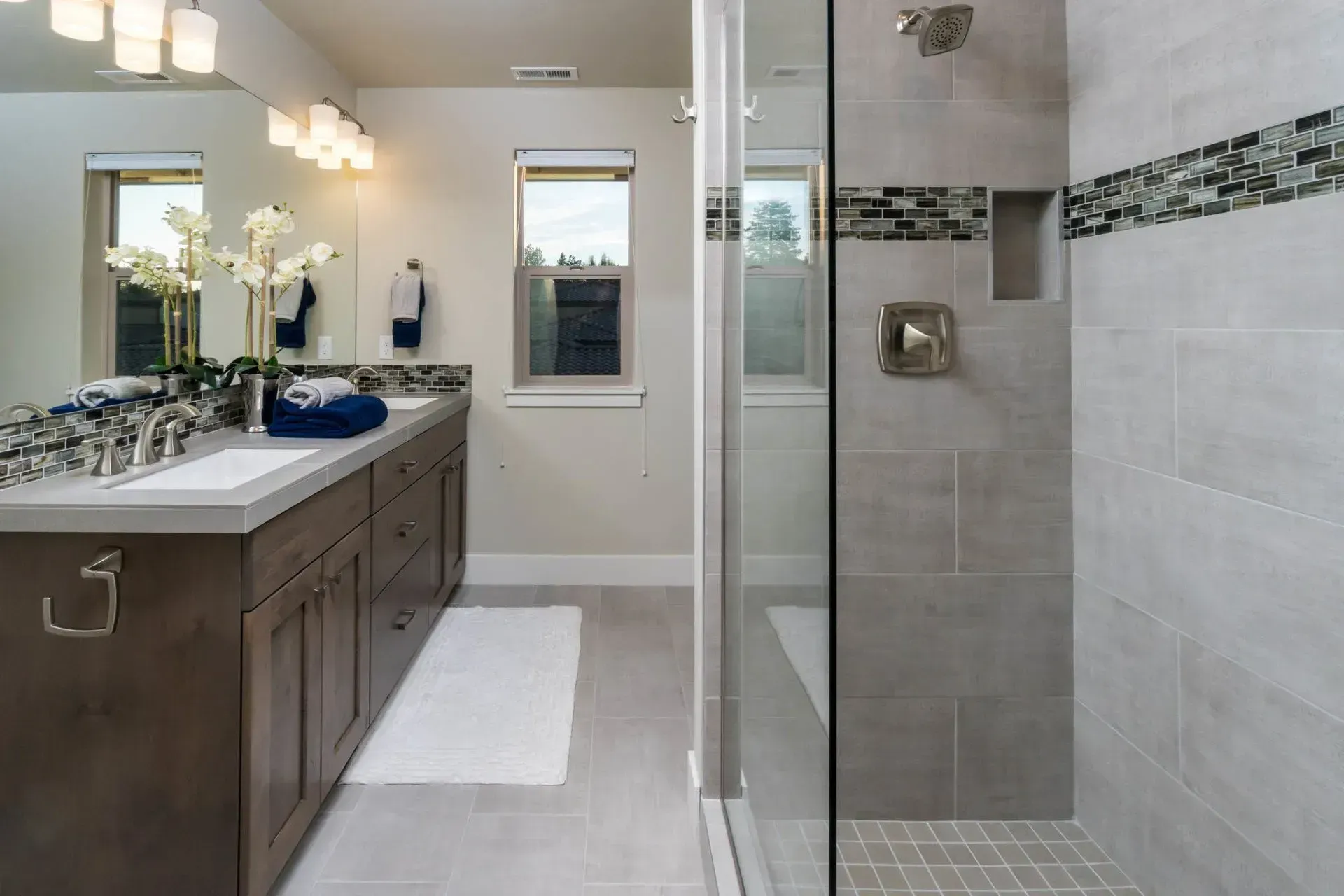Modern bathroom with double vanity, large mirror, and glass-enclosed shower in gray tile