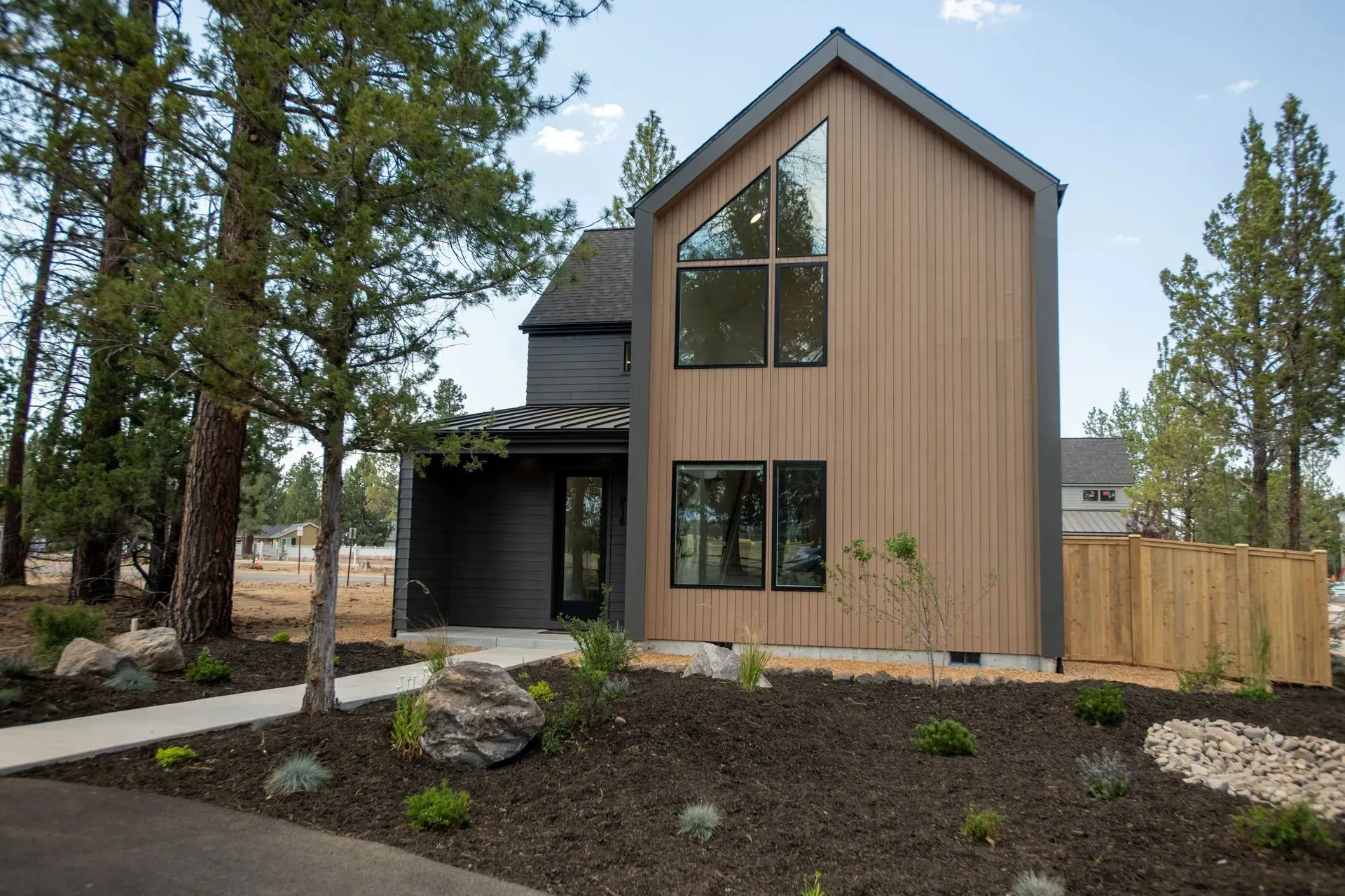 Modern two-story house with black trim, wood siding, and a landscaped front yard surrounded by tall pine trees
