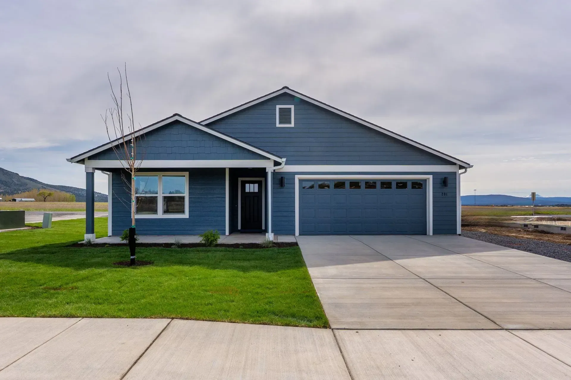 Blue single-story house with a two-car garage, front lawn, and wide concrete driveway under cloudy skies