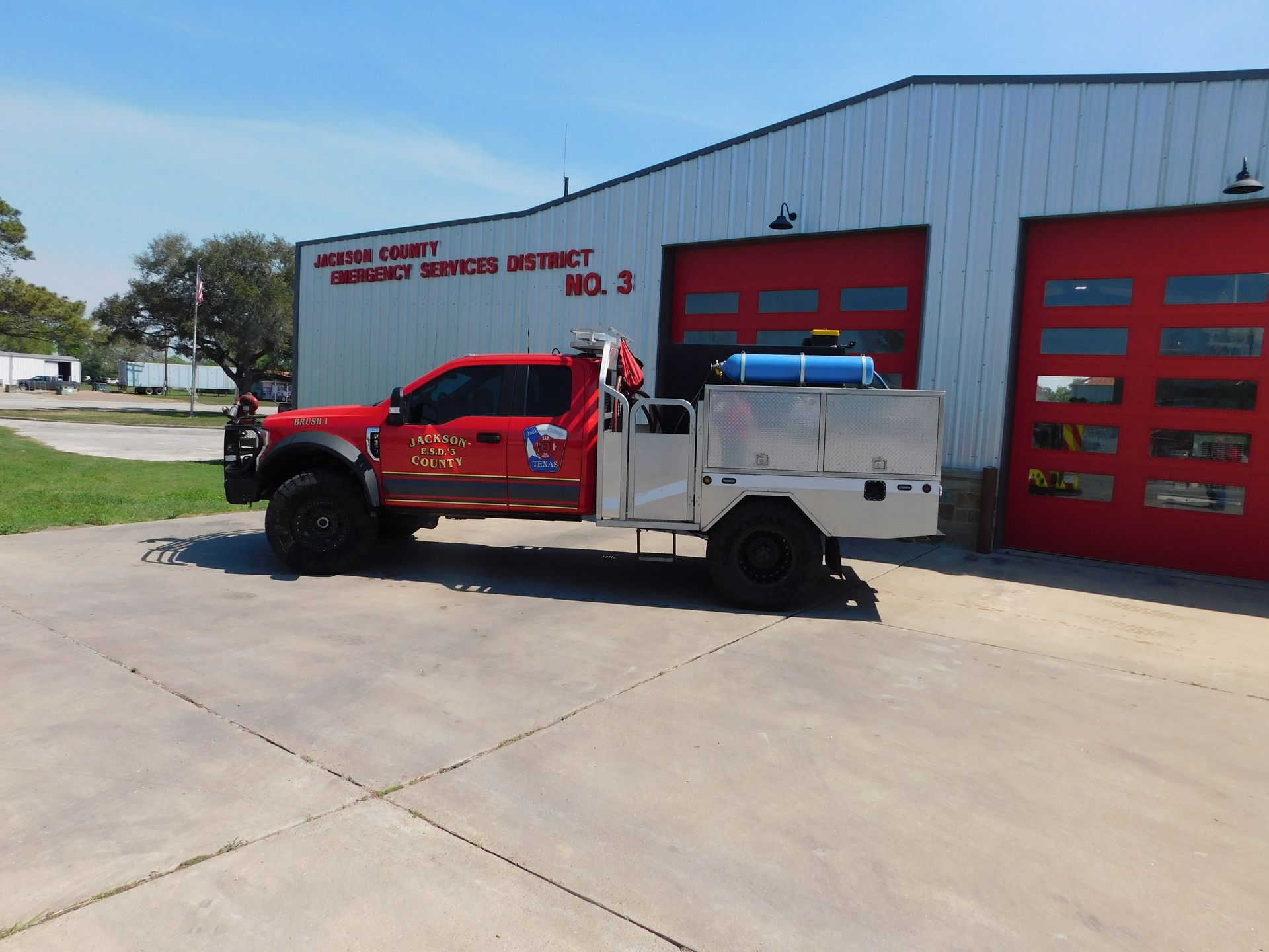 A side view of a red fire department brush truck parked on a grassy field under a sunny blue sky.