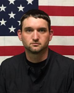A person with short dark hair in a black collared shirt stands in front of a United States flag.