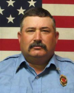 A man with a mustache in a light blue uniform shirt with a badge, positioned in front of an American flag.