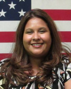 A smiling person with long, wavy brown hair, wearing a patterned top in front of an American flag background.