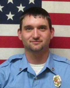 A person in a light blue uniform with a badge stands in front of a U.S. flag.