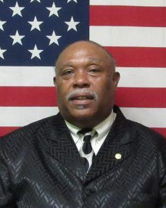 A person wearing a patterned black suit and a white collared shirt with a tie, posed in front of an American flag.