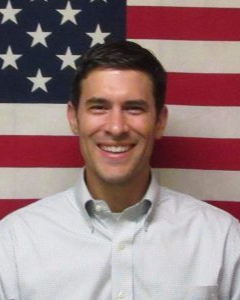 A smiling person wearing a button-down shirt, positioned in front of an American flag background.