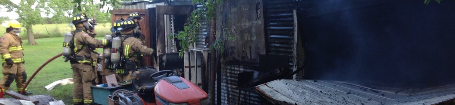 Firefighters in full protective gear use a hose to extinguish a fire in a charred wooden structure.