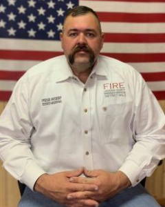 A man with a mustache, wearing a white button-down shirt with a fire district logo, sits before an American flag.