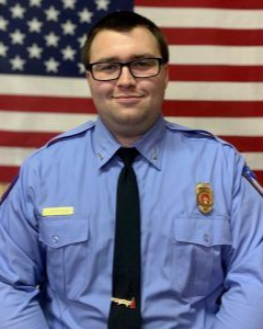 A person in a light blue uniform with a badge and black tie stands in front of an American flag.